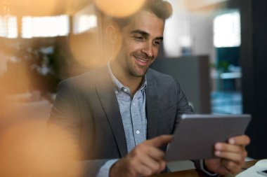 Gülümseyen genç işadamı sessiz bir ofiste masasında oturuyor ve dijital tabletle internette çalışıyor.