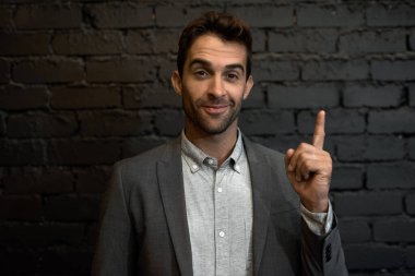 Genç bir işadamının portresi, bir ofiste koyu tuğla bir duvarın önünde dikilirken yukarı doğru işaret ediyor.