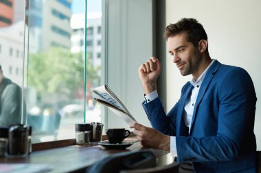 Genç işadamı kahve içip gazete okurken bir kafedeki pencerenin yanında oturuyor.