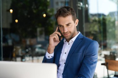 Genç işadamı dışarıda bir kafe masasında otururken cep telefonuyla konuşuyor ve dizüstü bilgisayarla çalışıyor.