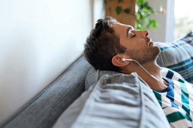 Genç adam evdeki kanepesinde oturuyor ve kulaklıkla gözleri kapalı müzik dinliyor.