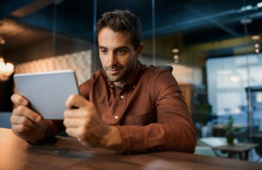 İş adamı gecenin erken saatlerinde karanlık ofis masasında tek başına oturuyor ve dijital tabletle internette çalışıyor.