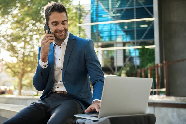 Smiling Young Businessman Using Laptop Talking His Cellphone While Sitting — Stock Photo, Image