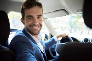 Genç bir adamın portresi omuzlarının üzerinden bakıyor ve bir arabanın sürücü koltuğunda otururken gülümsüyor.