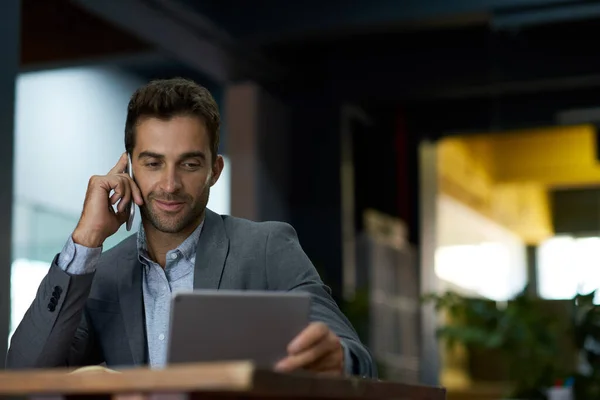 年轻的商人在一个安静的办公室里 一边用手机聊天 一边在办公桌旁做着一个数字平板电脑 — 图库照片