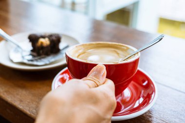 Ahşap bir masanın üzerinde kırmızı bir fincan kahveyle bir kadının elini kapat. Arka planda brownili beyaz bir tabak var.