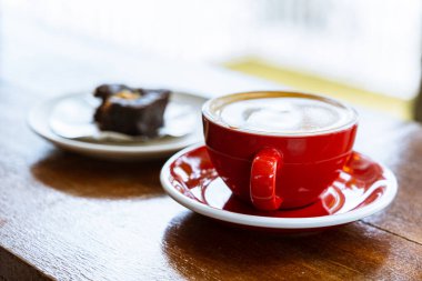 Ahşap bir masanın üzerindeki kırmızı bir fincan kahveyi kapat. Arka planda brownili beyaz bir tabak var.
