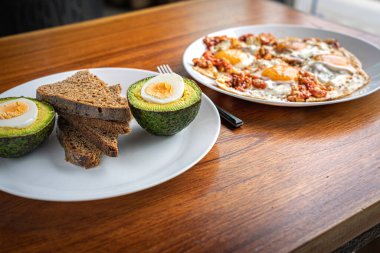 İkiye bölünmüş taze avokado, bir dilim ekmek ve ahşap masada beyaz tabakta domatesli sahanda yumurta.