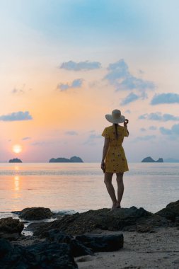 Kumlu sahilde şapkalı ve sarı elbiseli genç beyaz kadın günbatımının tadını çıkarıyor.