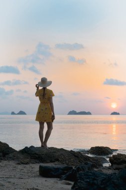 Güneş batarken kumlu sahilde şapkalı ve sarı elbiseli beyaz bir kadın batan güneşe bakıyor.