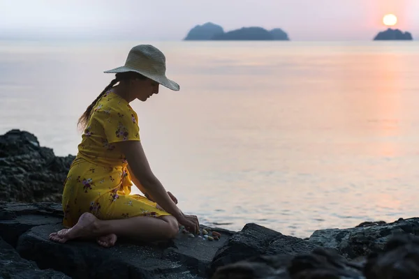戴着帽子和黄色衣服的年轻的高加索女人坐在海滩上的石头上 日落时与小石块和沙子玩耍 夕阳西下 — 图库照片