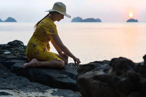 戴着帽子和黄色衣服的年轻的高加索女人坐在海滩上的石头上 日落时与小石块和沙子玩耍 夕阳西下 — 图库照片