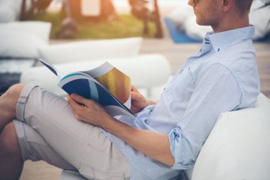 Kafkasyalı genç adam kanepe okuyucusunda dergi okuyor elinde bir kitap, açık bir sayfa tutuyor ve açık bir şekilde kütüphanede kanepede oturmuş kitap okuyor. Erkekler için yaşam tarzı.