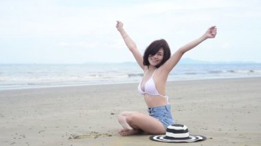 Yaz zamanı kadın bikinisi sahilde tatil yapıyor. Neşeli kadın sahilde oturmuş denize bakarken yazlık elbiseler ve hasır şapkalar giyiyor. Yazın tropikal ada sahillerinde dinlenme zamanı..