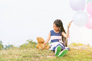 Mutlu Çocuk kucaklaşması oyuncak ayı yeşil parkta balon tutuyor. Oyuncak ayı, küçük çocuklar için en iyi arkadaş tatlı kız. Otizm, oyun alanında renkli helyum balonları tutarak birlikte çalmaktan mutluluk duyar.