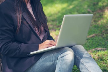 Asyalı kadın yeşil parkta dizüstü bilgisayar kullanarak oturuyor. Dizüstü bilgisayarlı mutlu girişimci işi yapan kadın Coronavirus karantina döneminde elleri klavye ev ofisinde yazan bir not defteri kullanıyor.
