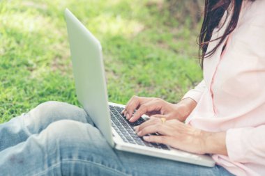 Asyalı kadın yeşil parkta dizüstü bilgisayar kullanarak oturuyor. Dizüstü bilgisayarlı mutlu girişimci işi yapan kadın Coronavirus karantina döneminde elleri klavye ev ofisinde yazan bir not defteri kullanıyor.