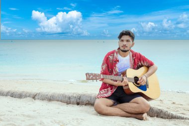 Genç erkekler plajda akustik gitar çalıyorlar. Akustik gitar enstrümanı çalan mutlu bir adam kumsalda, tropik plaj adasında oturuyor. Asyalı müzisyen Mutlu gitarist hobi yaşam tarzı