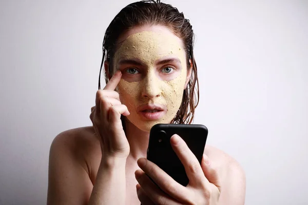 Retrato de una joven mujer caucásica con máscara de arcilla facial usando su teléfono inteligente viendo videos. Espacio para texto . —  Fotos de Stock
