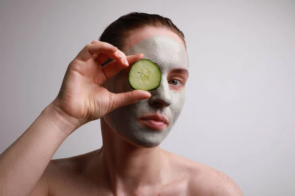 Mujer joven con una mascarilla en la celebración de un rodajas de pepino delante de un ojo. Concepto de belleza y cuidado de la piel . —  Fotos de Stock