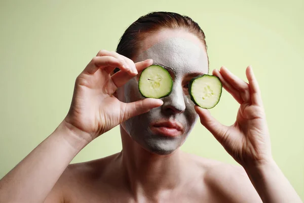 Arcmaszkos fiatal nő portréja, amint egy uborka szeletet tart az egyik szeme előtt. Szépség és bőr koncepció. — Stock Fotó