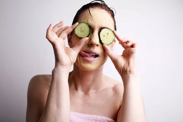 Mujer joven feliz con una mascarilla en la celebración de un rodajas de pepino en frente de los ojos. Concepto de belleza y cuidado de la piel . —  Fotos de Stock