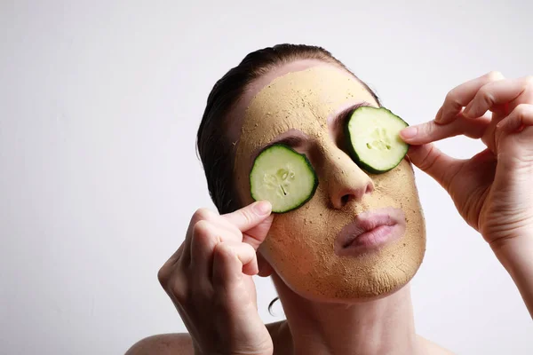 Young woman with a face mask on holding a cucumber slices in front of one eye. Beauty and skincare concept. — Stock Photo, Image
