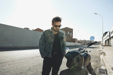 Kot ceketli ve güneş gözlüklü bir adam tozlu toprak yolda yarış motosikleti sürüyor.. 