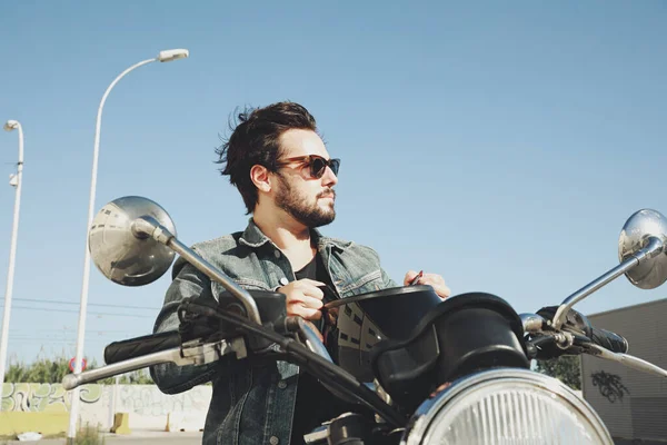 Sakallı mutlu motosikletçi elinde kaskla yolda duruyor..