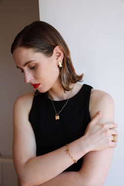 Güzel bir kadının dikey portresi kırmızı dudaklarıyla yana bakıyor ve beyaz duvarın üzerinde altın takılar takıyor..