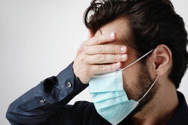 Tıbbi maskeli genç bir adam, beyaz arka planda duruyor ve alnına dokunuyor. Salgın durumu, Covid-19..