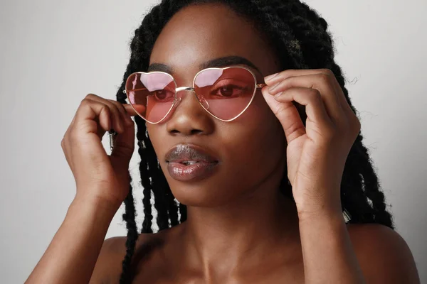 Mujer negra joven con trenzas de llegada con gafas de sol de color rosa, posando sobre fondo blanco. Concepto de cuidado de la piel. —  Fotos de Stock