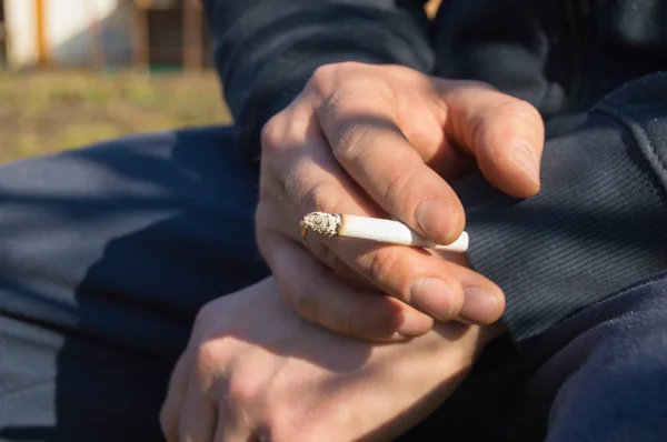 Молодий чоловік тримає цигарку в руці, сидячи на відкритому повітрі — стокове фото