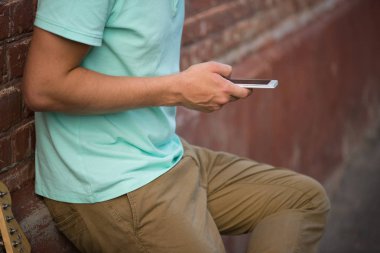 Genç şık adam kahverengi tuğla duvar, cep telefonu yanında gitar holding eller olmalıdır