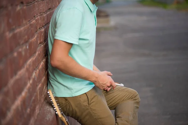 Joven chico con estilo debe tener una pared de ladrillo marrón, manos sosteniendo el teléfono celular al lado de la guitarra — Foto de Stock