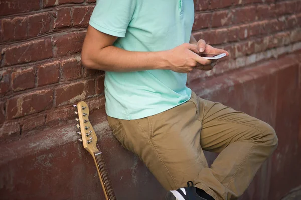 Jeune homme élégant devrait avoir un mur de briques brunes, les mains tenant le téléphone cellulaire à côté de la guitare — Photo