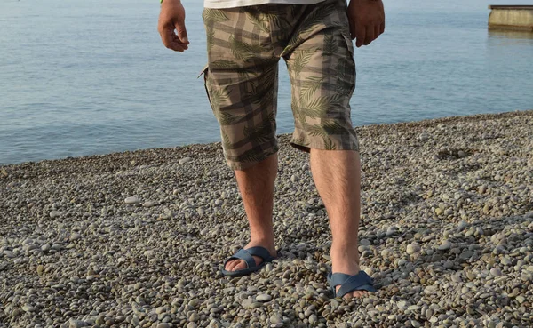 mens legs in flip-flops and shorts, a man standing on the beach on a pebble beach