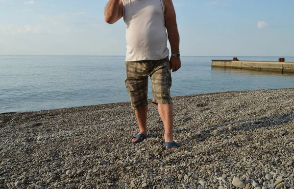 mens legs in flip-flops and shorts, a man standing on the beach on a pebble beach