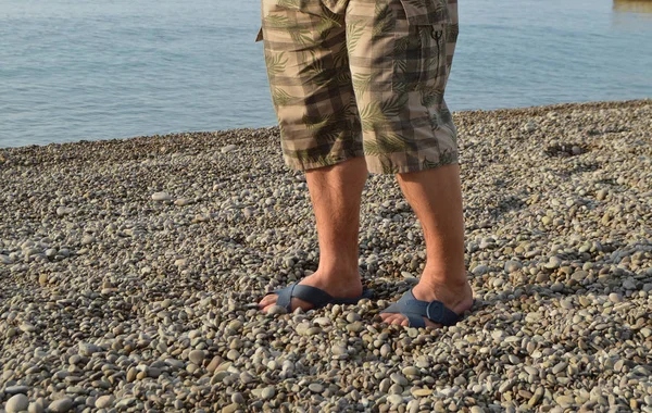 Мужские ноги в шлепанцах и шортах, мужчина, стоящий на пляже на галечном пляже — стоковое фото