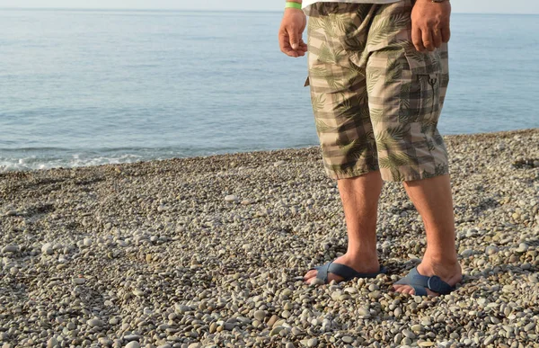 Mens bacaklarda Flip-flop ve şort, bir adam sahilde bir çakıl plajı üzerinde ayakta