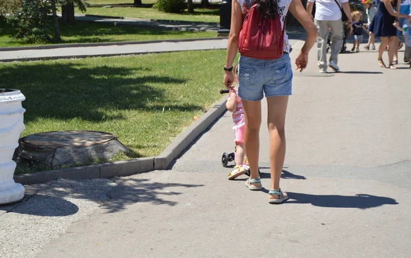 Malá roztomilá holčička, procházky v parku s skútr, vedle mladé matky v šortkách, s batohem — Stock fotografie