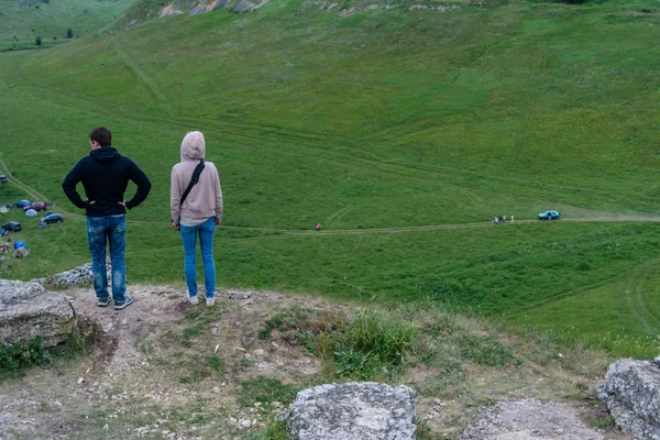 Rear view of a young couple, a sporty guy and a slender girl, stand on top of a mountain and look down at the camp of tourists. Tourism, the success and the concept of a healthy lifestyle. — Stock Photo, Image