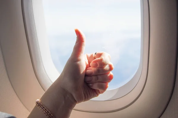 Gesture thumb up, OK sign, woman's hand on the background of the WINDOW on the plane.