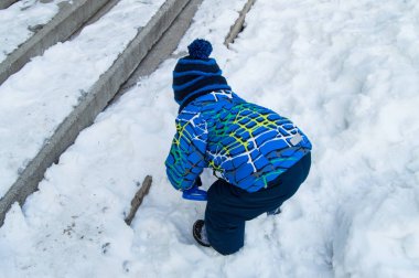 Mavi örgü şapkalı ve ceketli küçük bir çocuk kışın merdivenlerin yanında oynar. Karla kaplı, arkadan manzaralı..