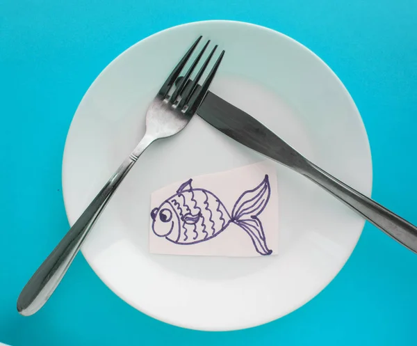 Sărbătorirea Zilei Prostului din aprilie, o farfurie cu furculiță și cuțit și un pește de hârtie pe un fundal albastru. Umor — Fotografie, imagine de stoc