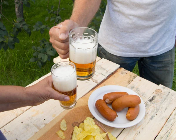 hands holding glasses with beer, chips and sausages lying on a light wooden boards, people relax in the weekends, Octoberfest