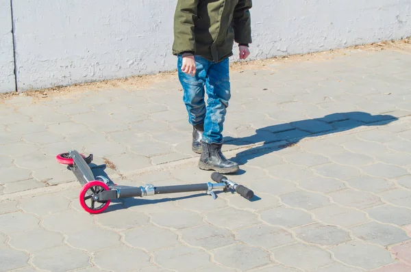 Niño adolescente con una chaqueta verde y jeans levanta su scooter después de una caída al aire libre en el parque, entretenimiento infantil al aire libre —  Fotos de Stock