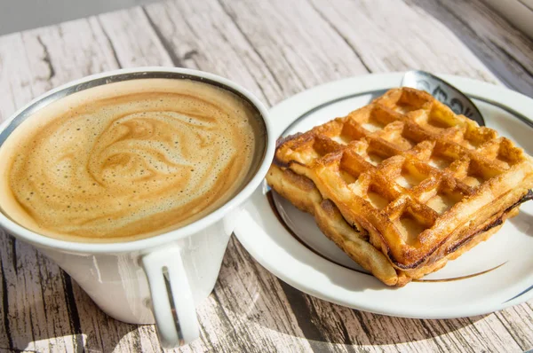 Ontbijt in zonlicht met vintage houten achtergrond, witte porseleinen beker met warme aromatische koffie met schuim en Weense wafel op een bord — Stockfoto