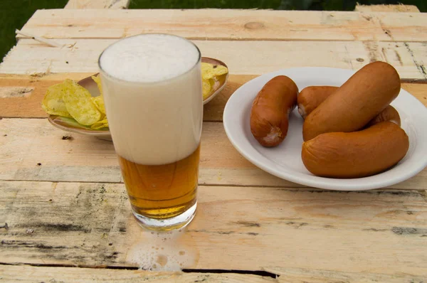 Cerveza en un vaso con espuma, salchichas, patatas fritas en las tablas del jardín, tema Oktoberfest . —  Fotos de Stock