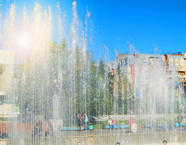 Fuente de la ciudad con chorros de agua y personas al azar, vista a través de salpicaduras en el día soleado de verano —  Fotos de Stock